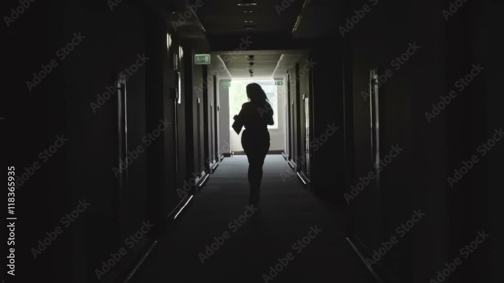 Backlit silhouette of woman with long fly away hair running alone through dark corridor in office building and holding file Backlit silhouette of woman with long fly away hair running alone through dark corridor in office building and holding file