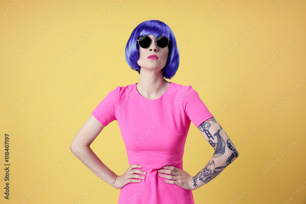 Beautiful young woman with tattoo wearing blue wig and sunglasses on yellow background Beautiful young woman with tattoo wearing blue wig and sunglasses on yellow background