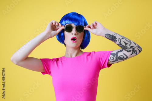 Beautiful young woman with tattoo wearing blue wig and sunglasses on yellow background