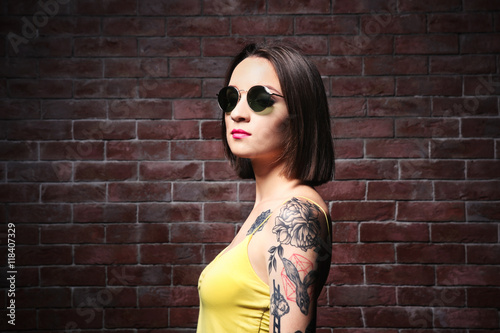 Beautiful young woman with tattoo wearing sunglasses and posing on brick wall background