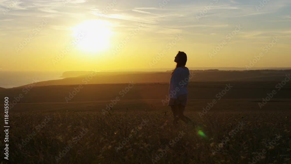Wide shot of young woman walking through a field at sunset by herself Wide shot of young woman walking through a field at sunset by herself