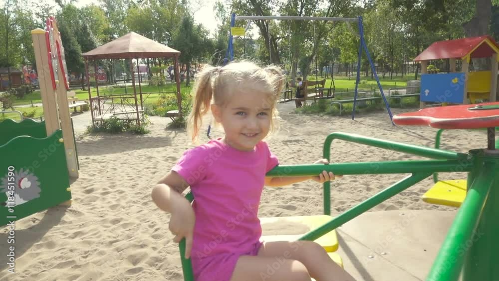 Adorable little girl  on the carousel  smiles for the camera. Child playing in the park sunny summer day. Slow motion.
