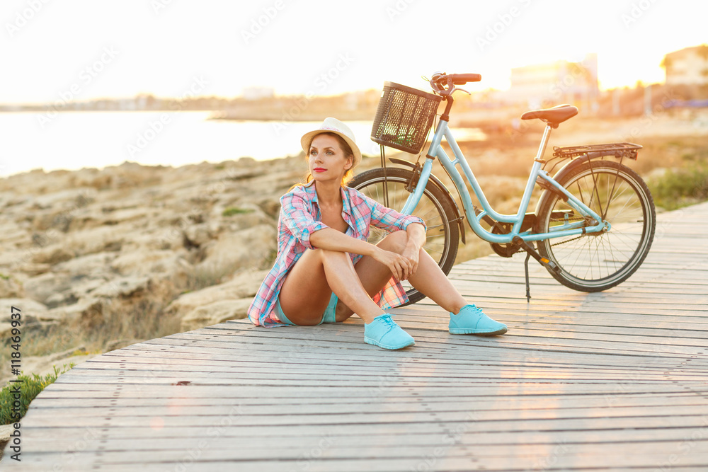 Carefree pretty woman with bicycle sitting on a wooden path at t
