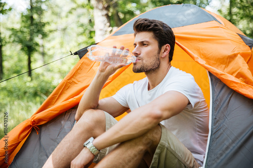 Man sitting near touristic tent and drinking water in forest