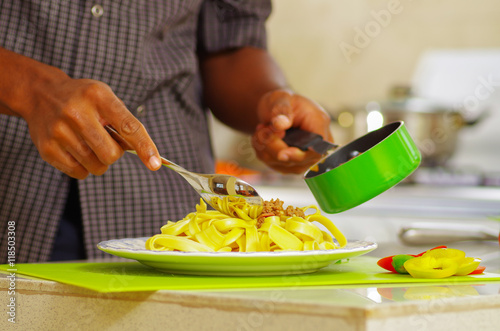 Closeup of mans hands transferring tagliatelle pasta fom green pot onto white plate, kitchen background