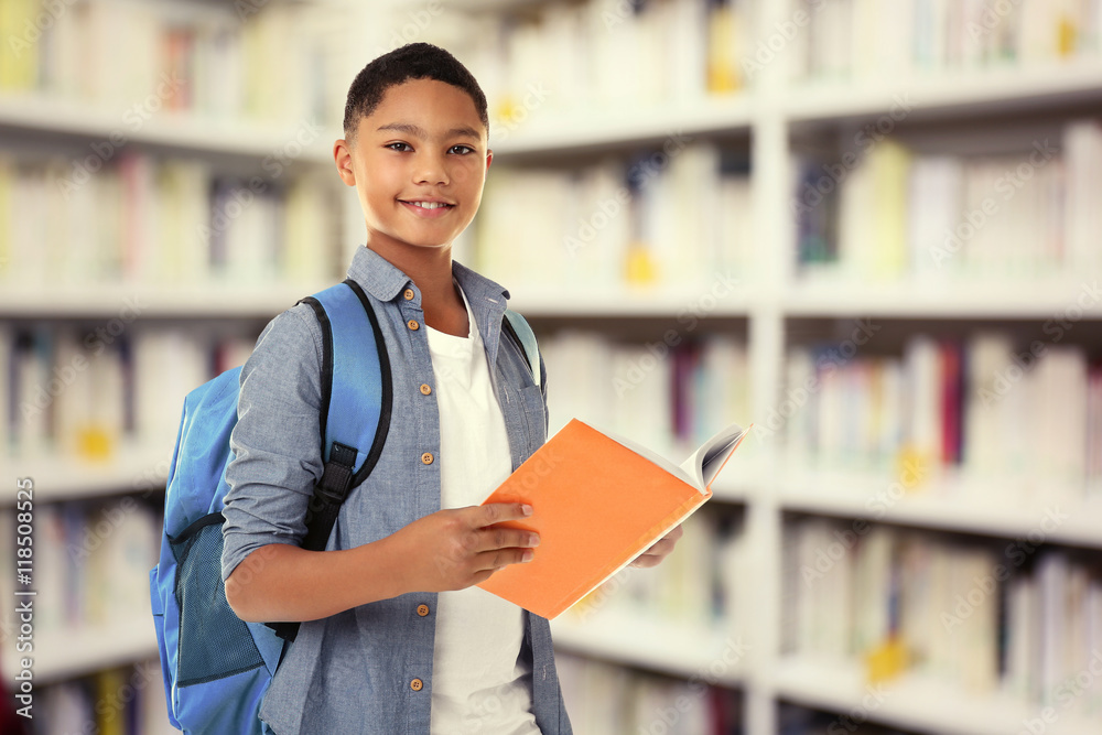 Cute boy with backpack holding book on blurred book shelves background. Library concept. Cute boy with backpack holding book on blurred book shelves background. Library concept.