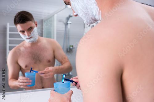 Handsome male with shaving foam on his cheeks washes his razor i