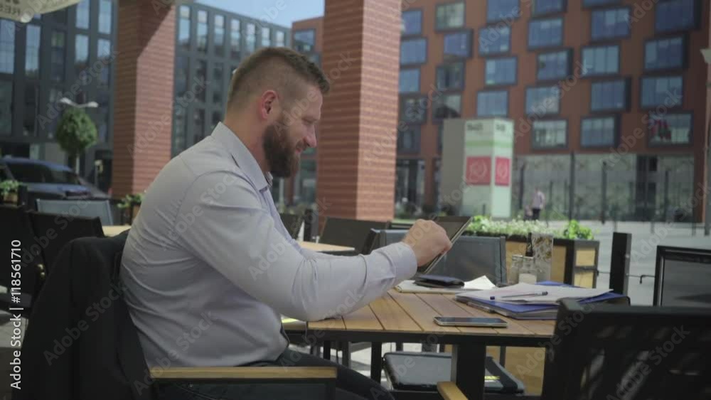 Happy and content businessman browsing tablet. He is sitting at the table in cafe. He is young and has beard. Man is dressed in suit and shirt. Happy and content businessman browsing tablet. He is sitting at the table in cafe. He is young and has beard. Man is dressed in suit and shirt.