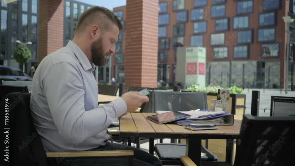 Businessman copies data from credit card to tablet. He is sitting at the table in cafe. He is young and has beard. Man is dressed in suit and shirt. Businessman copies data from credit card to tablet. He is sitting at the table in cafe. He is young and has beard. Man is dressed in suit and shirt.