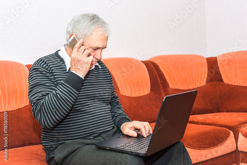 Senior businessman talking on cell phone and typing on laptop sittin on sofa. Concept photo of senior people and modern technology.