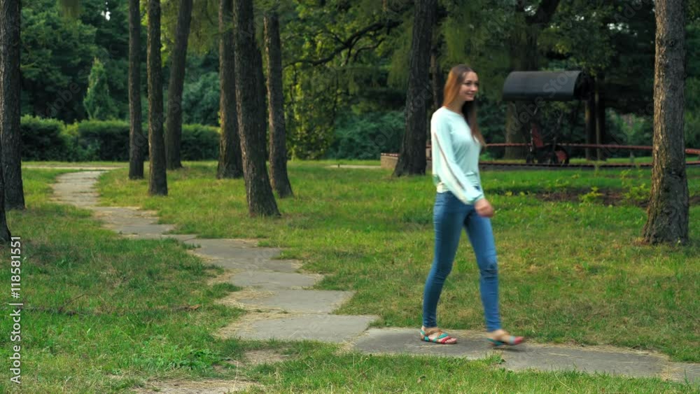 Young brunette girl with long hair in bright sandals is on a path in a park and glances up at the trees and the sun. Young brunette girl with long hair in bright sandals is on a path in a park and glances up at the trees and the sun.