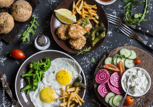 Lunch table - fried eggs, fish balls, potato chips, vegetables, sauces, homemade bread on a dark background. Rustic style, top view