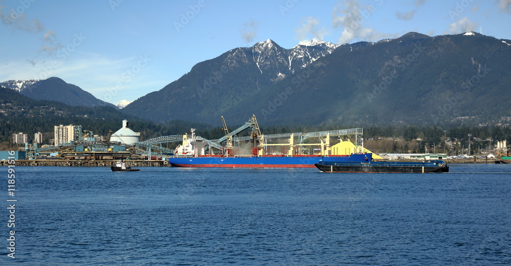 Terminal of North Vancouver port on a background of mountain scenery. The vessel is under load, the tug is towing another vessel.