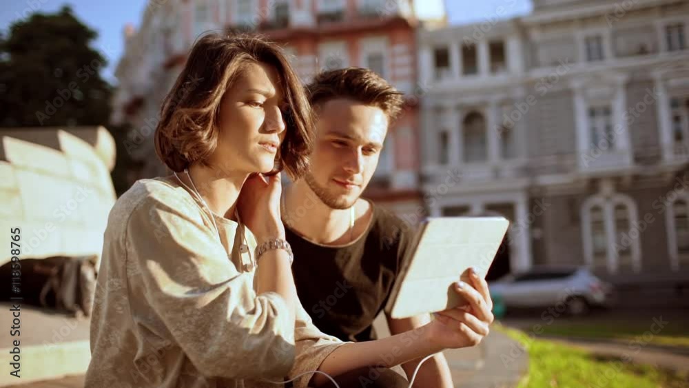 Young beautiful couple smiling, speaking, looking at tablet, sitting in city park. Slow motion. Young beautiful couple smiling, speaking, looking at tablet, sitting in city park. Slow motion.