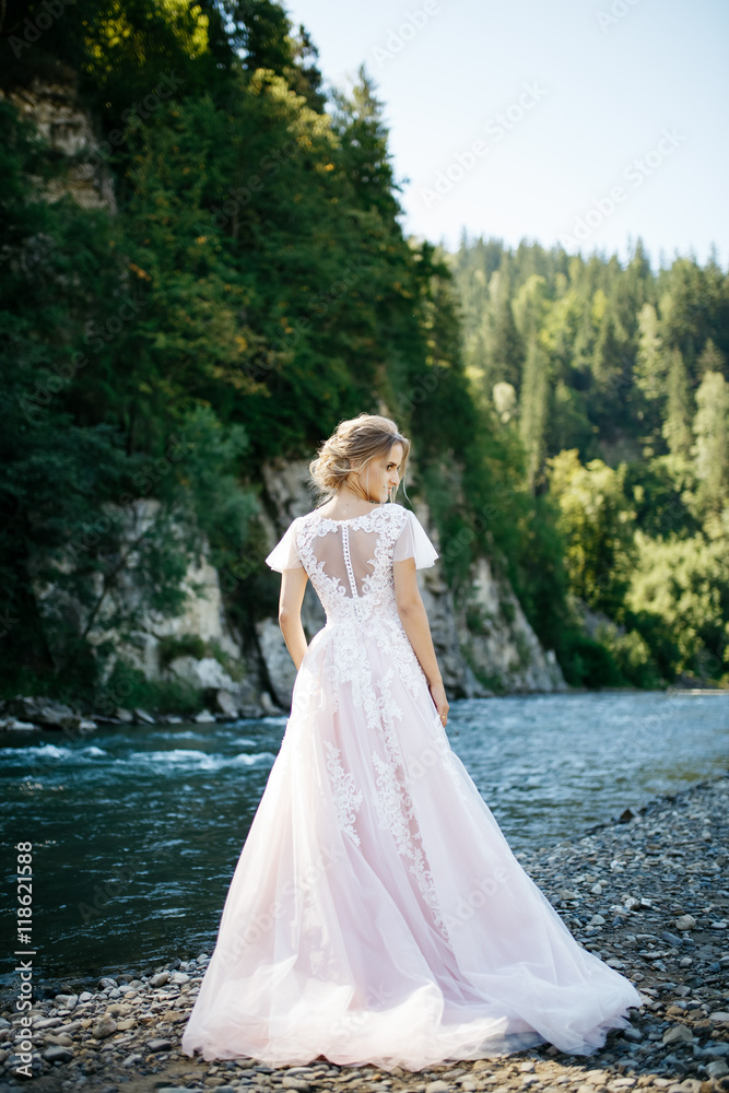 Blonde bride stands in the rays of sun on river's shore