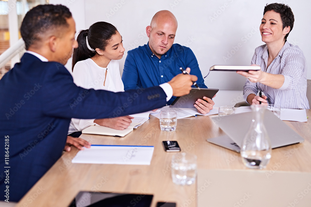 Four business partners together in a conference room with meeting underway as mature caucasian female passed her mixed race business partner seated on the other side of the table. Four business partners together in a conference room with meeting underway as mature caucasian female passed her mixed race business partner seated on the other side of the table.