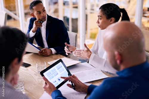 Diverse group of four business individuals listening to a young female partner busy talking to them while looking up the figures she is referring to on his electronic tablet in real time.