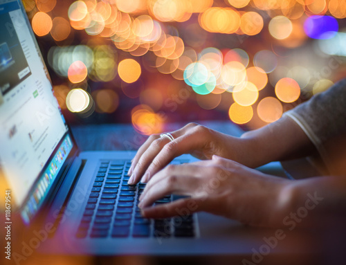 Woman hands typing on laptop keyboard on abstract blurred bokeh of city night light background. Focus in the foreground.