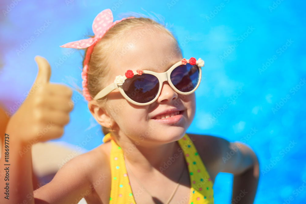 Little girl showing thumbs up on the pool background. Happy smiling Child is wearing sunglasses, enjoying pool and having fun. Travel (vacation), adventure concept. Close up, outdoor. Little girl showing thumbs up on the pool background. Happy smiling Child is wearing sunglasses, enjoying pool and having fun. Travel (vacation), adventure concept. Close up, outdoor.