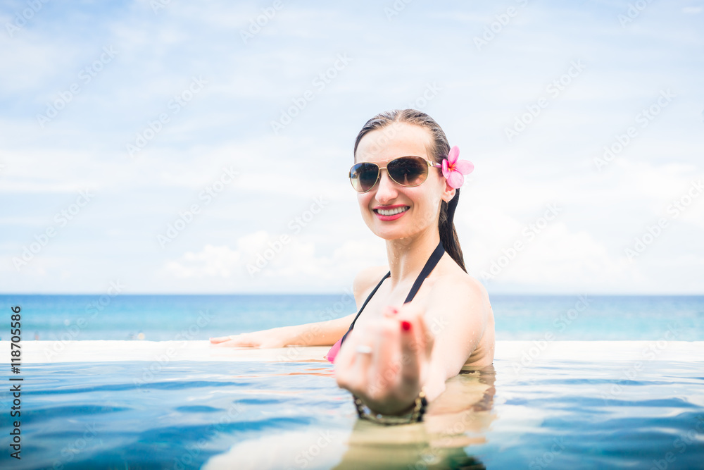 Frau mit Sonnenbrille macht lockende Geste am Rand von Infinity-Pool