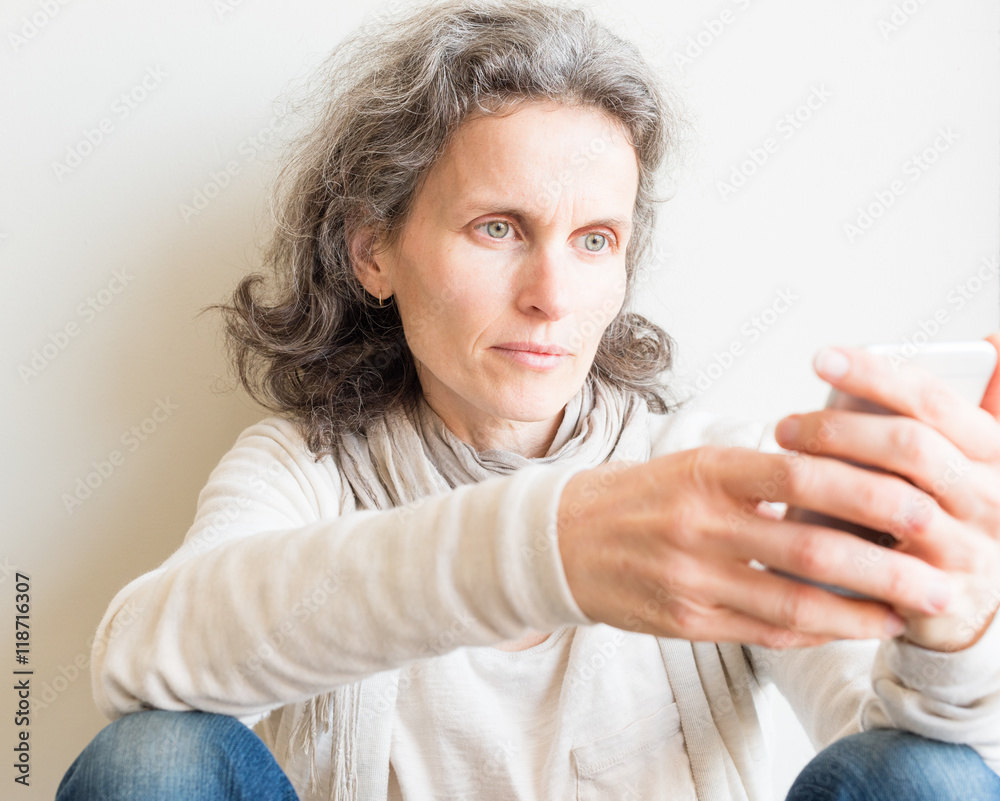 Middle aged woman with grey hair and cream top sitting on floor and looking intently at smart phone (selective focus and cropped) Middle aged woman with grey hair and cream top sitting on floor and looking intently at smart phone (selective focus and cropped)
