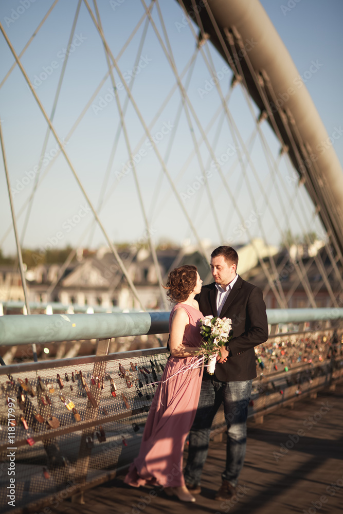 Fototapeta premium Stylish loving wedding couple, groom, bride with pink dress kissing and hugging on a bridge at sunset