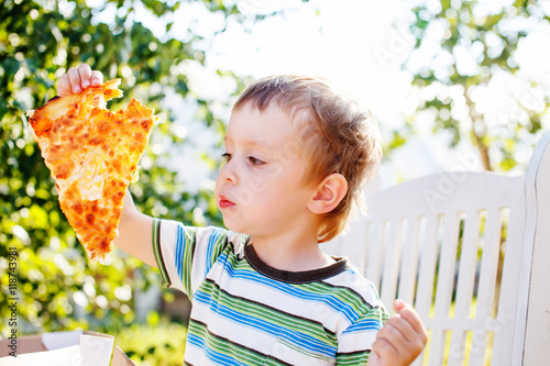 kid enjoying pizza outdoors. boy holding a slice of pizza aweigh and intending to bite off photo