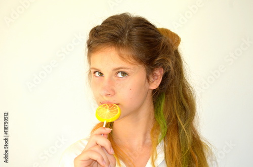 Молодая  девушка с разноцветными волосами  в белой рубашке на белом фоне с леденцом в руках в виде  лимона 
