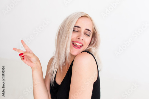 Portrait of cheerful woman with peace hand sign. Drunk playful blonde in black dress with closed eyes laughing and posing jocosely at camera, white background