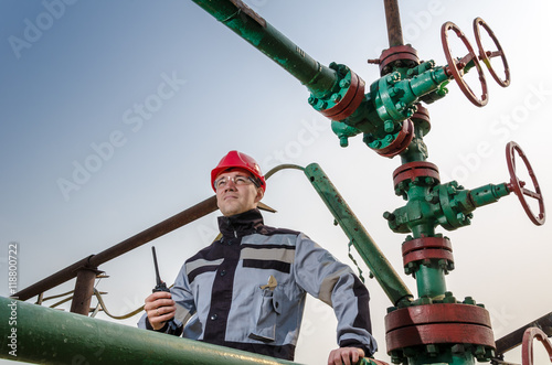 Oilfield worker near wellhead valve wearing red helmet and work clothes talking on the radio. Oil and gas concept.