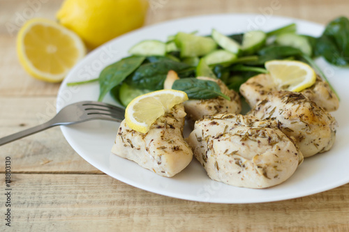 Baked chicken breast served on a white plate with lemon, baby spinach and fresh cucumber