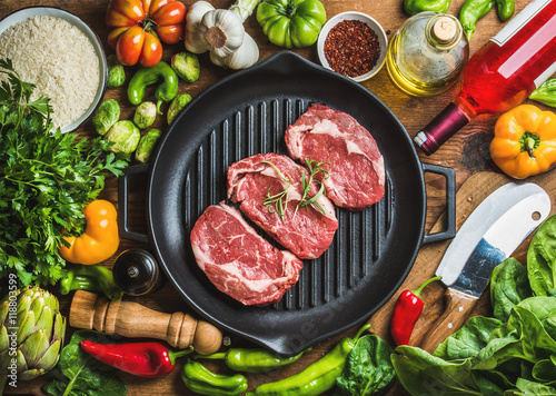 Ingredients for cooking healthy meat dinner. Raw uncooked beef steaks in cast iron grilling pan with vegetables, rice, herbs, spices and rose wine bottle over wooden background, top view