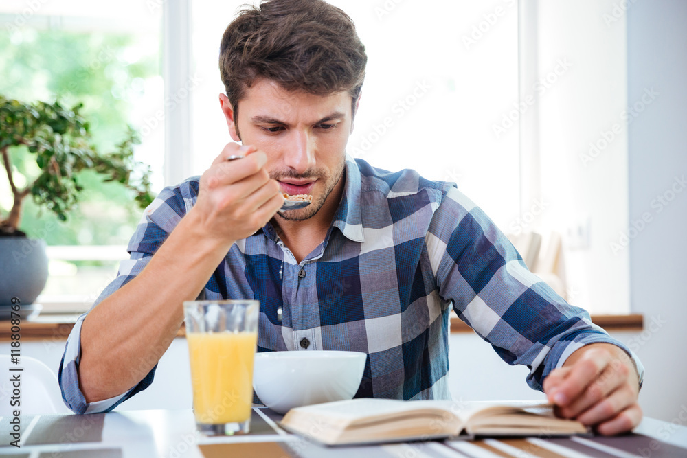 Concentrated young man reading book and having breakfast on kitchen Concentrated young man reading book and having breakfast on kitchen