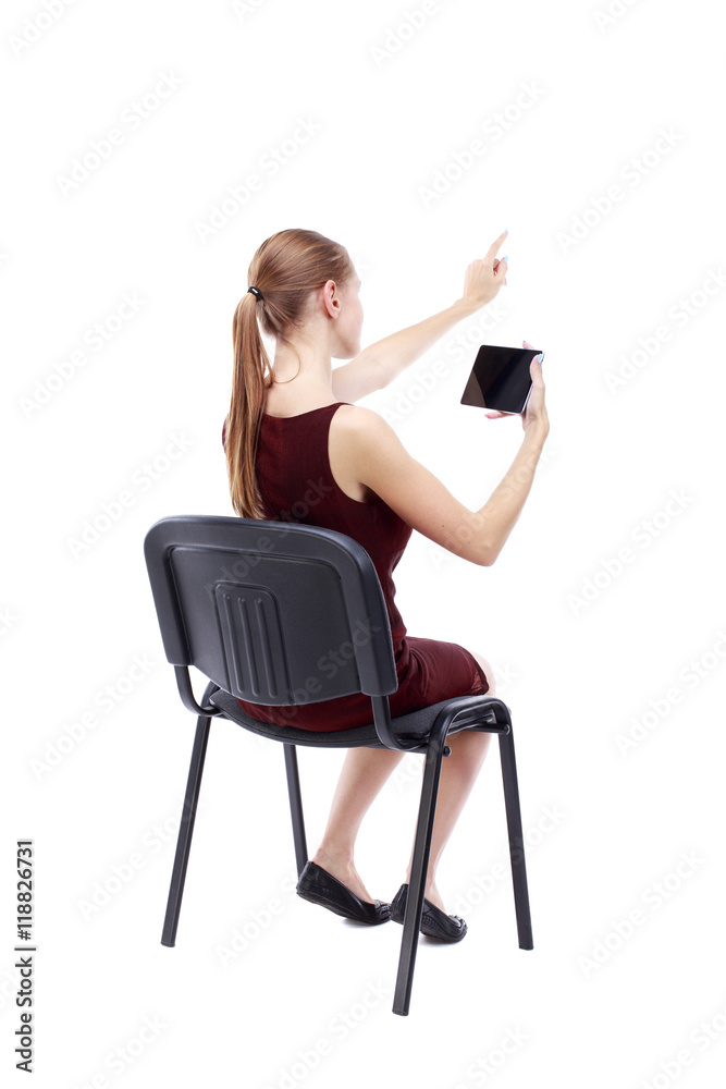 back view of pointing woman sitting on chair and looks at the screen of the tablet.  Rear view people collection.  backside view of person.  Isolated over white background. A girl in a burgundy dress
