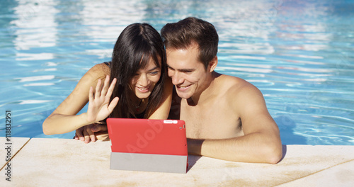 Couple communicates with friends on digital device with red cover while in swimming pool