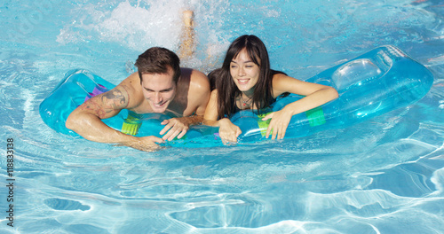 Pair of joyful male and female adults swimming together on inflatable floating plastic mattress in outdoor pool
