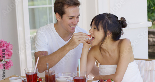 Smiling man feeding his wife bits of fruit at breakfast with pink flowers and waffles in front of them