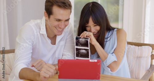 Blushing couple showing off ultrasound pictures to someone through a video chat on their tablet computer © Dash