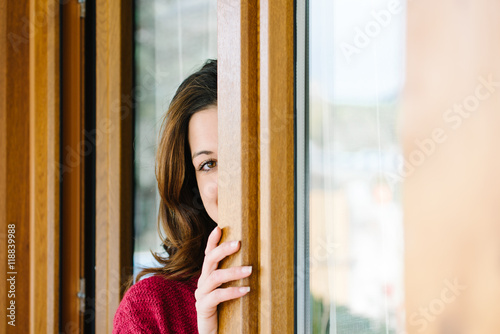 Playful young woman having fun peeking through the window of her home. Adult girl hiding for looking and watching outside.