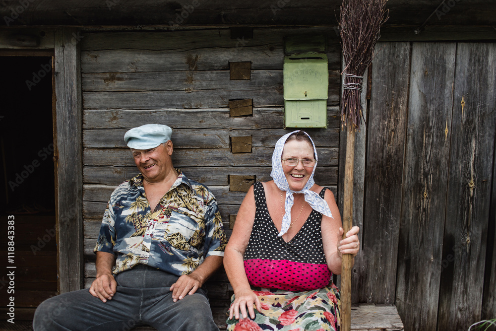 beautiful mature russian couple have fun outside of their house in village. The woman clothed in a scarf keeps the broom