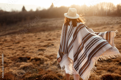 Fotografia happy boho woman, walking towards sun, wearing hat and poncho at