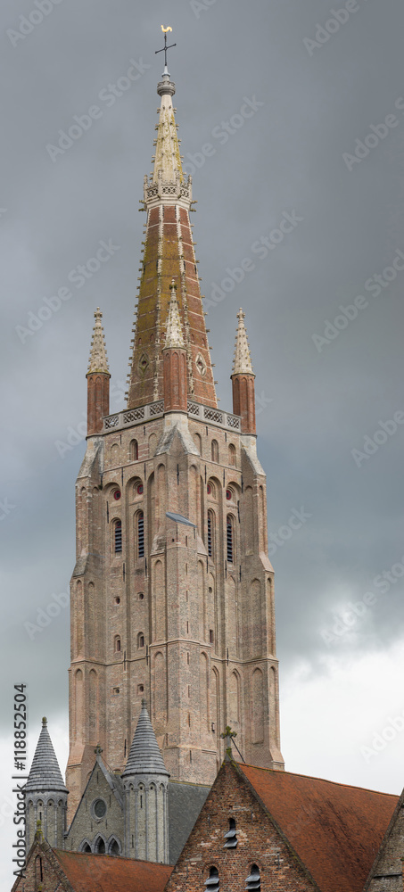 Brugge, Belgium - August 10, 2016: The brick tower of Notre Dame Cathedral, Onze-Lieve-Vrouw, stands against stormy rainy dark sky. Old Sint Jans Hospital in front.