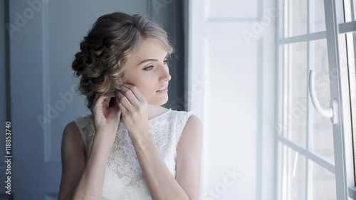 Beautiful bride in white wedding dress puts on earring. Beauty model girl is wearing jewelry. Female portrait in bridal gown for marriage. Woman with curly hair and lace veil. Cute lady indoors