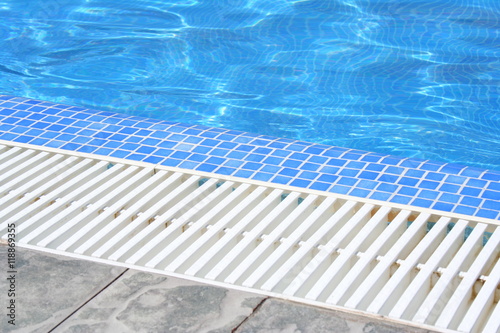 Swimming pool white grating grille with clear blue water and grey tiles.