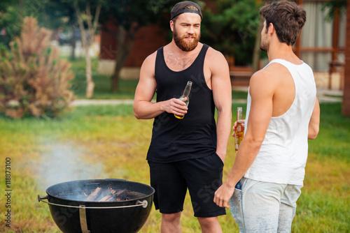 Two men holding a beer bottle while preparing barbecue