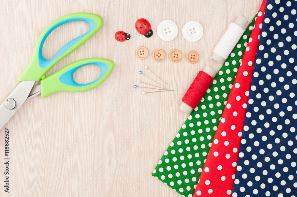 Fototapeta premium Fabric for sewing and accessories for needlework on wooden background. Spool of thread, scissors, buttons, sewing supplies