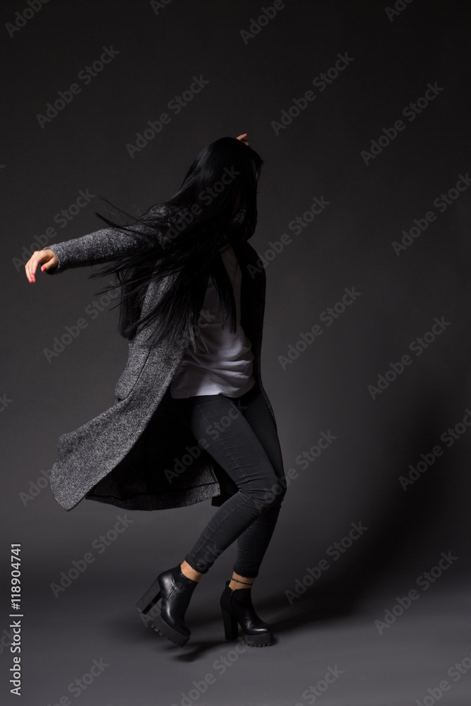 Brunette model woman dancing in studio while posing for photographer demonstrating designers' clothes isolated on grey background.