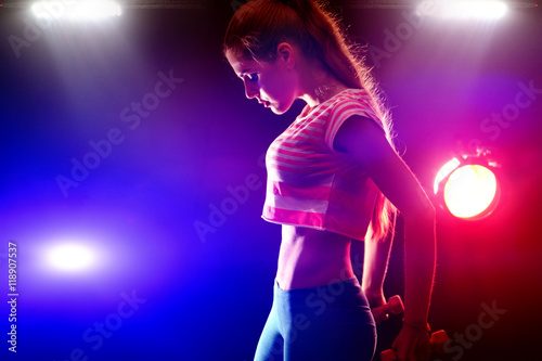Sports fitness girl with slim stomach works dumbbells in multi-colored rays of the fitness room.