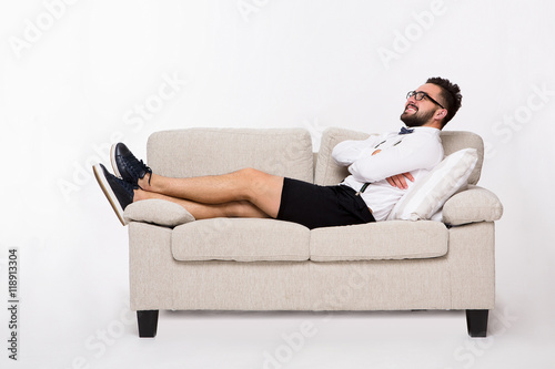 Picture of smiling handsome hipster man lying on couch or sofa and looking upwards. Young man lying with his arms crossed or folded isolated on white background. © Svyatoslav Lypynskyy