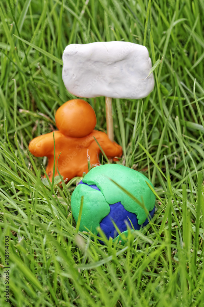 Fototapeta premium Plasticine globe in tall grass. A modelling clay man holds a placard. The concept of Earth Day.
